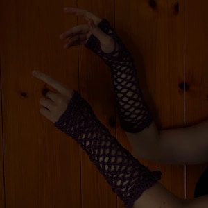 May include: Dark purple, open-weave arm warmers. The crochet gloves have a scalloped edge at the wrist and a fingerless design. The arm warmers are displayed against a wooden background.