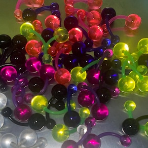 May include: A selection of brightly coloured belly button rings in shades of pink, red, green, yellow, purple, and black. The curved bars and round balls create a lively display. These body jewellery pieces are made of clear, translucent material.