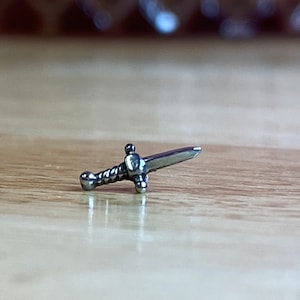 May include: A silver-toned sword charm with a twisted handle and a pointed tip. The charm is lying on a wooden surface.