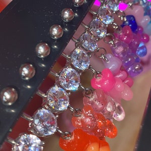 May include: A set of colorful gummy bear charm earrings with clear rhinestone accents. Each earring features a different color gummy bear charm, including yellow, orange, red, pink, purple, and blue.