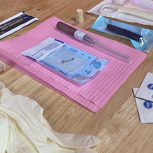 May include: A pink sterile surgical pad with a clear plastic tube, a small silver piercing, a pair of white tweezers, a pair of blue tweezers, a black marker, a cork stopper, a pair of white latex gloves, and two small white packets with the word "Kroger" printed on them.
