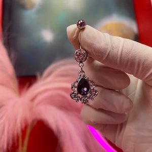 May include: A silver belly button ring with a dangling purple gem and ornate silver design.
