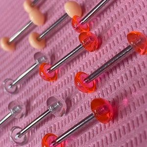 Puede incluir: Primer plano de varios pares de joyas para el cuerpo. Las joyas están hechas de acrílico transparente y rosa y presentan un estilo de barra con un extremo de tornillo.