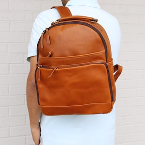 May include: A brown leather backpack with multiple zippered compartments and a top handle. The backpack has adjustable shoulder straps and a rounded top. The bag is worn over the shoulder.