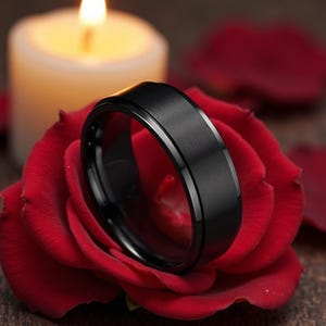 以下が含まれることがあります： 赤バラの上に置かれた、つや消し加工された金属の中央バンドと磨かれたエッジを持つ黒い指輪。背景には、散らばったバラの花びらとともに、火の灯った白いろうそくがあります。