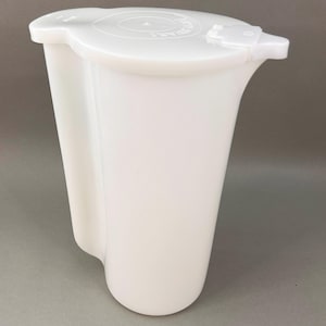 May include: A white plastic pitcher with a tall, cylindrical body and a flat, circular lid. The lid features a small, hinged spout cover and embossed text that reads Tupperware. The pitcher has a molded handle integrated into the side wall.