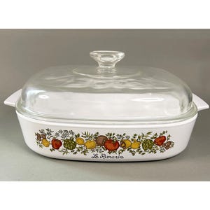 May include: White casserole dish with a clear glass lid and handle. The dish features a colorful border of fruits and vegetables. The words "Le Romarin" are printed on the front.