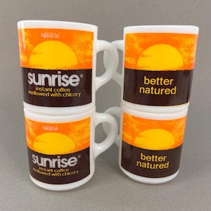 May include: Four white ceramic coffee mugs stacked. Each mug features a sunrise design in orange and yellow, with the word "sunrise" or "better natured" in brown. The mugs have a handle and the Nestle logo.