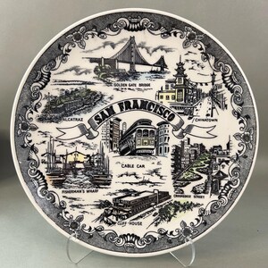 May include: A round white ceramic decorative plate featuring black and white illustrations of San Francisco landmarks including the Golden Gate Bridge, Alcatraz, Chinatown, a cable car, Fisherman's Wharf, and the Cliff House with floral borders.