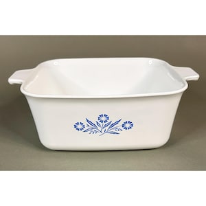 May include: A square white ceramic baking dish with integrated side handles. The exterior features a centered blue floral pattern consisting of three stylized flowers with pointed leaves. The interior is smooth, white, and deep with rounded corners.