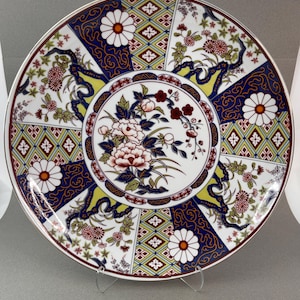 May include: A decorative ceramic plate with a detailed floral design. The plate features a central floral arrangement surrounded by a border of alternating blue, white, and yellow sections with floral and geometric patterns. The plate has a gold rim.