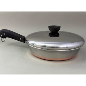 May include: Stainless steel frying pan with a lid. The pan has a copper-colored base and a black handle. The lid has a black knob. The pan is designed for cooking.