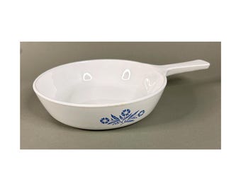 May include: White oval serving dish with a handle. The dish features a blue floral pattern on the side. The handle is rectangular and extends from the side of the dish. The dish is made of ceramic.
