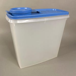 May include: A clear, rectangular food storage container with a blue lid and a small, round, blue lid on top. The container is made of a translucent plastic material.