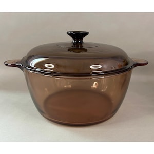May include: A round, translucent amber-coloured glass casserole dish with a matching lid. The dish features two integrated side handles and a central knob on the lid. The glass is smooth and tinted brown, allowing light to pass through the material.