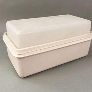 May include: A rectangular, off-white plastic storage container with a matching lid. The container features a smooth, matte finish and a simple, functional design. A small, circular embossed logo is visible on the center of the lid surface.