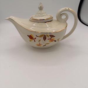 May include: A cream-coloured ceramic teapot featuring a gold-trimmed rim and lid. The body is decorated with a floral pattern in shades of brown, orange, and yellow. It has a curved handle with a circular cutout and a pointed spout.