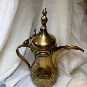 May include: A vintage brass coffee pot with a long spout and a curved handle. The pot has an ornate design on the lower portion and a decorative finial on the lid. The brass has a warm, golden hue.