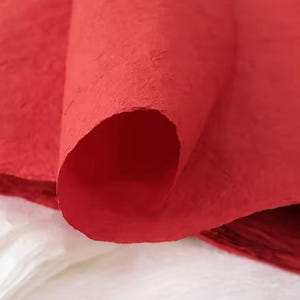 May include: Close-up of a red textured paper sheet. The paper is rolled up and the texture is visible. The paper is a deep red color.