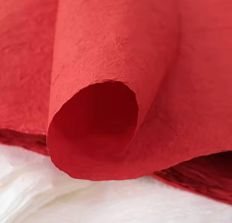 May include: Close-up of a red textured paper sheet. The paper is rolled up and the texture is visible. The paper is a deep red color.