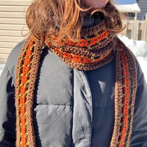 Peut inclure: Écharpe au crochet faite à la main, aux tons marron, orange et vert. L'écharpe est enroulée autour du cou. Le motif est texturé et réalisé en fil doux. L'adresse du site web est visible en bas. Un accessoire d'hiver confortable.