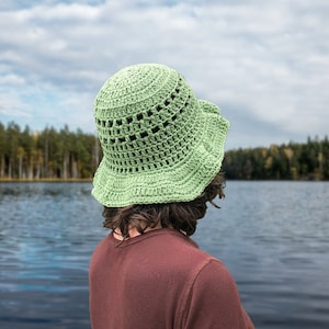 May include: A person wearing a green crocheted sun hat with a wide brim, standing with their back to the camera, looking out at a lake. The lake is surrounded by trees and the sky is cloudy.