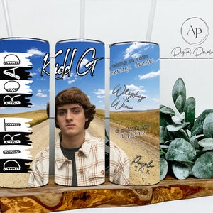 May include: Three stainless steel tumblers with different designs. The first tumbler has a blue sky background with white clouds and the text "Dirt Road" in black. The second tumbler has a photo of a young man with the text "Kidd G" in black. The third tumbler has a blue sky background with white clouds and the text "Could be your teenage dream" in black. The text "Whiskey & Wine" is also on the tumbler in black. The text "If heaven had a phone" is on the tumbler in black. The text "People Talk" is on the tumbler in black.