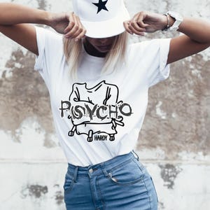 May include: White t-shirt with the word "PSYCHO" in a distressed font and a black graphic. The shirt is paired with blue jeans and a white baseball cap with black star accents. The cap is worn with the brim facing forward.