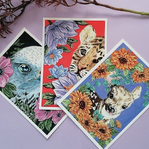 Könnte beinhalten: Drei Kunstdrucke mit Tierporträts und Blumenrändern. Einer zeigt einen Vogel mit rosa Blumen, ein anderer einen Tiger mit lila Blüten und der letzte ein Reh mit orangefarbenen Blumen. Jeder Druck ist rautenförmig und hat einen weißen Rand.