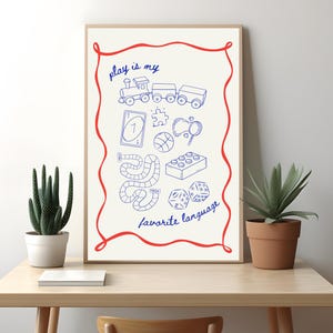 May include: A framed print with a cream background and a red border. The print features blue line drawings of toys and games, including a train, a playing card, a basketball, and dice. The text "play is my favorite language" is written in blue script.