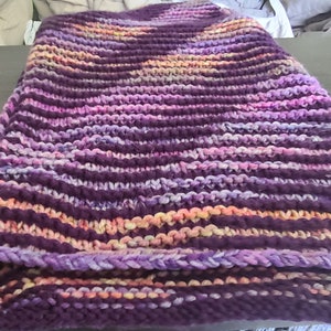 May include: A hand-knitted throw blanket in shades of purple, pink, and orange. The blanket is made with a chunky yarn and has a textured stitch pattern.