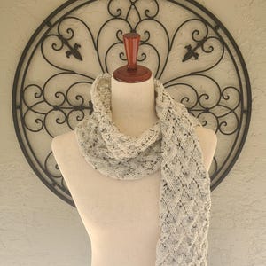 May include: A cream-colored knitted scarf with a delicate cable pattern. The scarf is draped over a white mannequin against a background of a wrought iron decorative wall.