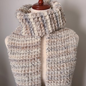 May include: A white, brown and grey knitted scarf with a textured pattern. The scarf is folded in half and draped over a mannequin.