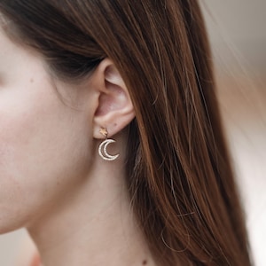 Puede incluir: Pendientes de oro en forma de media luna con un pequeño amuleto de estrella que cuelga de la parte inferior. Los pendientes se llevan en la oreja de una persona.