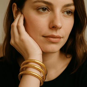 Peut inclure: Gros plan d'une personne portant trois bracelets dorés. Les bracelets sont épais, texturés et ont un aspect légèrement martelé. La personne porte un haut noir et a la main sur le visage.