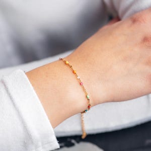Peut inclure: Un bracelet doré délicat orné de petites perles colorées. Porté au poignet, le bracelet présente des couleurs vives : orange, jaune, rose et vert. La chaîne réglable ajoute à son design élégant.