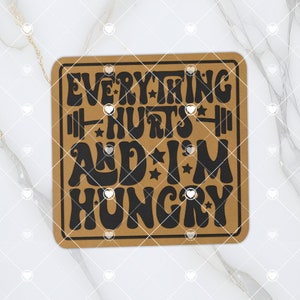 May include: A brown square coaster with the text "Everything Hurts And I'm Hungry" in black, stylized font with stars and a barbell graphic.