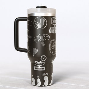 May include: Black travel mug with a white design of medical symbols and illustrations related to pregnancy and childbirth. The mug has a black handle and a silver lid.