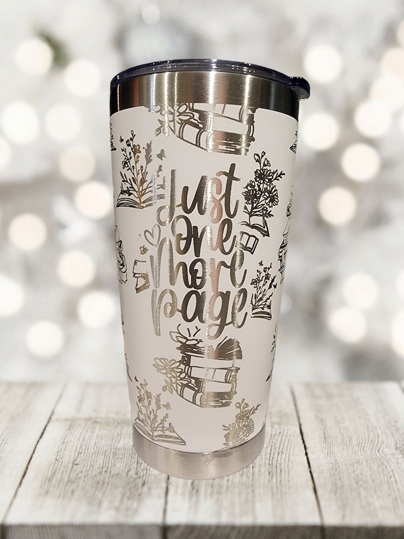 May include: White stainless steel tumbler with a silver design of books and flowers. The text "Just one more page" is printed on the tumbler.