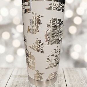 May include: White stainless steel tumbler with a silver metallic design of books, flowers, and moons.