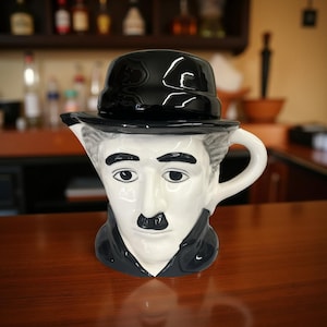 May include: A ceramic pitcher shaped like Charlie Chaplin's face, featuring a black bowler hat, a black mustache, and a white face. The pitcher has a handle and a spout, and is set on a wooden surface.