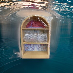 Puede incluir: Una estantería de madera con un letrero rojo y blanco que dice "The Christmas Palace". La estantería tiene tres estantes y está decorada con una escena navideña blanca y azul. La estantería está envuelta en plástico transparente.