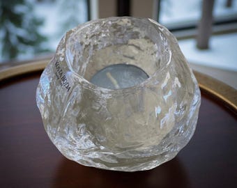 May include: A clear, textured glass votive candle holder with a teal candle inside. The candle holder has an organic, irregular shape and the text "KOSTA BODA" is visible on the side. The holder sits on a dark brown surface.