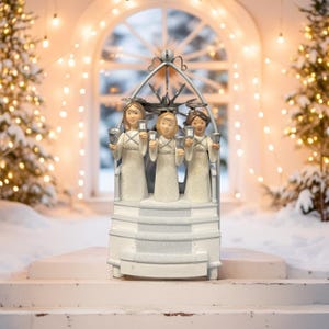May include: A white Christmas decoration featuring three angel figurines holding silver bells. The angels are inside a decorative arch, with a snowy backdrop and string lights. The overall design evokes a festive, winter holiday theme.
