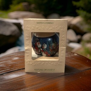 Pode incluir: Um enfeite de Natal vintage em uma caixa. O enfeite é uma bola de vidro redonda com fundo azul e uma cena de Papai Noel e outras figuras. A caixa é branca com o texto "Santa's Coming!" e "Keepsake Ornament".