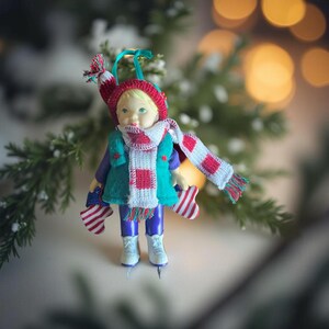 Peut inclure: Ornement de Noël représentant une figurine de poupée portant un bonnet, une écharpe et des mitaines rouges et blanches. La poupée porte une veste turquoise, une chemise violette et un pantalon bleu. La poupée porte des patins à glace blancs.