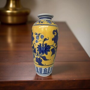 Pode incluir: Um vaso de cerâmica amarelo com desenhos florais azuis. O vaso tem um pescoço estreito e uma base larga. O padrão floral azul inclui flores e folhas. O vaso está sobre uma superfície de madeira escura.