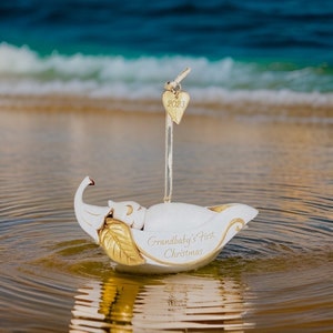 Op de afbeelding: Witte keramische ornament in de vorm van een slapende baby in een boot in de vorm van een blad. De ornament heeft gouden accenten en de tekst "Grandbaby's First Christmas" aan de zijkant. Een gouden hartbedel met het jaar 2023 is aan de bovenkant bevestigd.