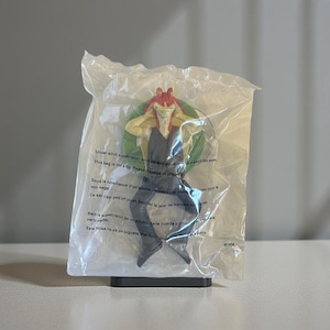 Peut inclure: Une figurine de Jar Jar Binks dans un sac en plastique transparent. La figurine porte une tenue noire avec des accents rouges et jaunes. Le sac contient du texte en plusieurs langues, dont le français.