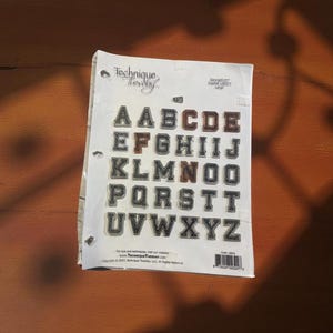 Op de afbeelding: Een vel alfabetletterstickers in collegestijl. De letters zijn in verschillende kleuren, waaronder bruin, zwart en wit. Het vel is gelabeld met "Technique Tuesday" en bevat het volledige alfabet.